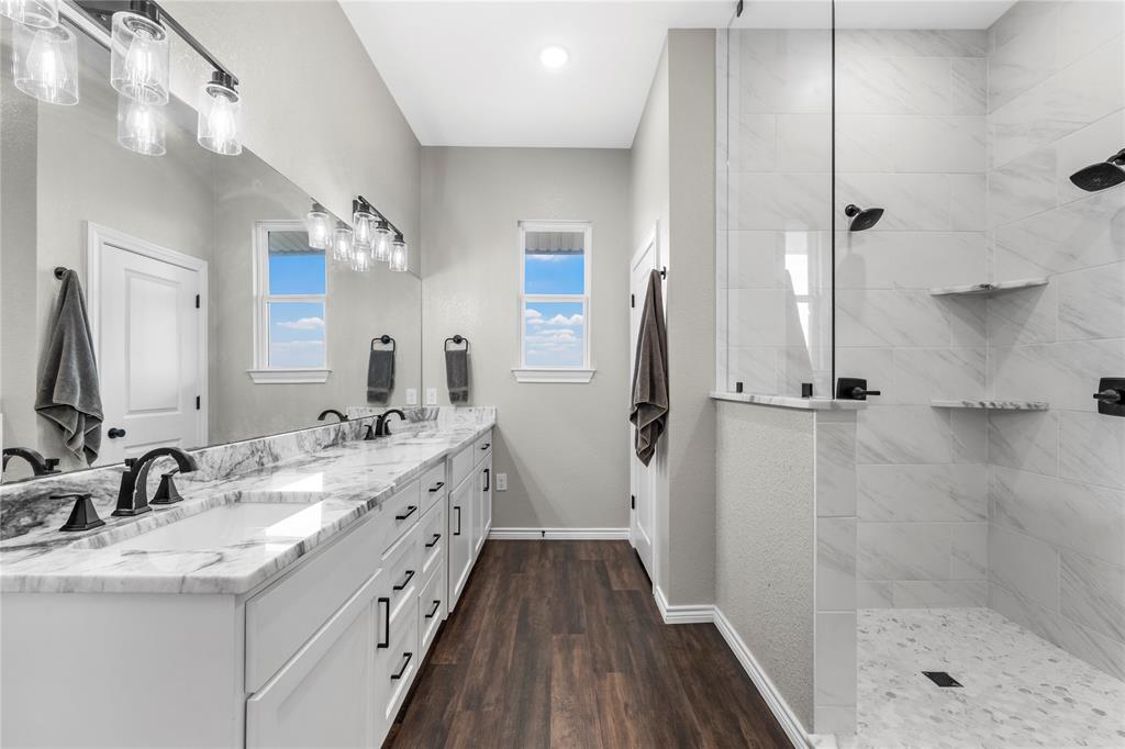 Dual vanity with stone-look countertop, walk-in shower featuring glass enclosure and stone-look tile, wood-finish flooring, and multi-light fixtures