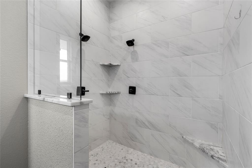 Walk-in shower featuring floor-to-ceiling rectified-edge tile, a frameless glass partition, matte black rainfall showerhead, dual built-in corner shelving, and a pebble-stone basin floor