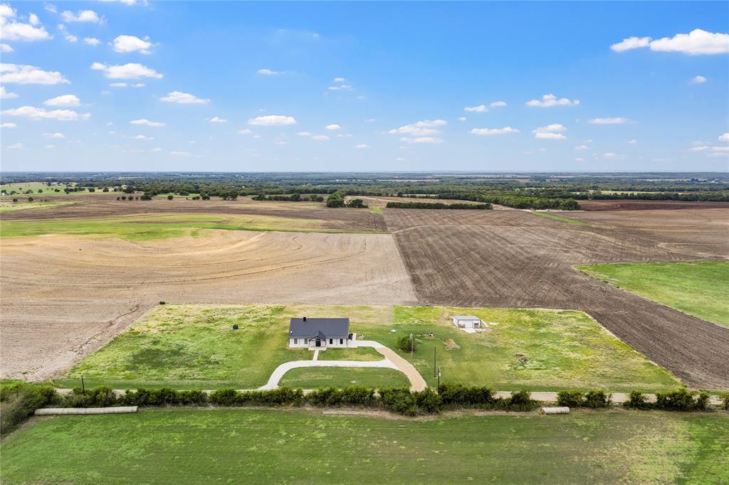 Aerial perspective featuring a single-story residence with a metal roof, a detached outbuilding, a gravel driveway, and surrounding acreage