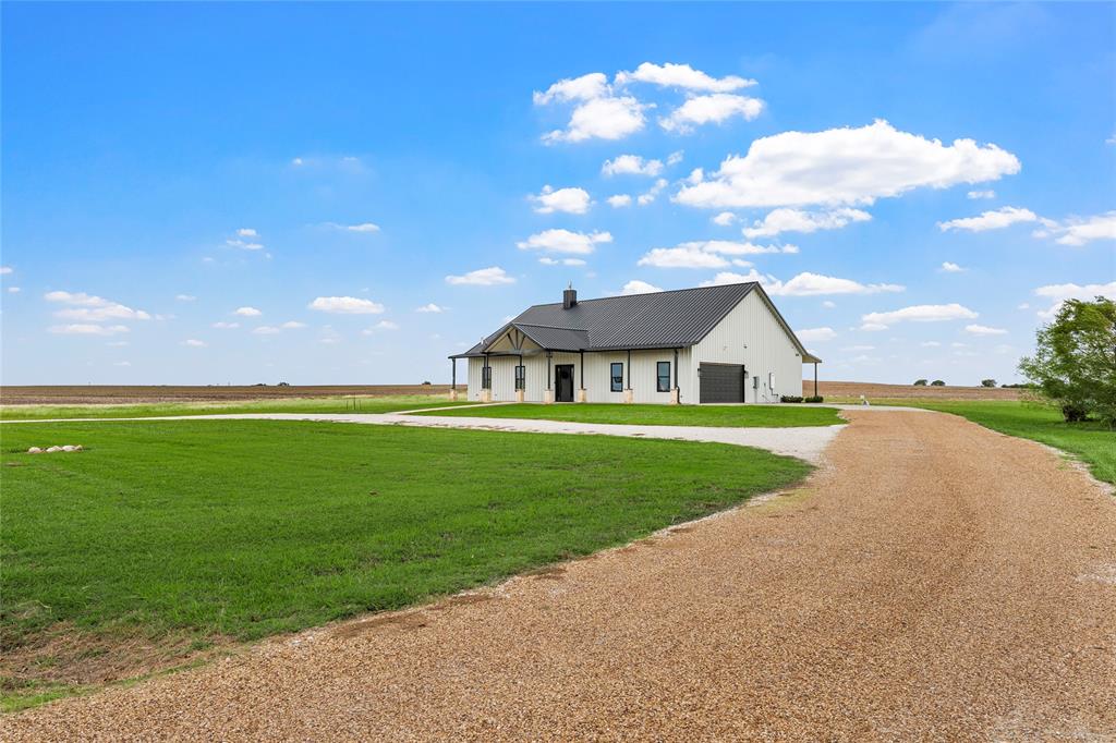 Single-story residence featuring a metal roof, light-toned exterior cladding, an attached garage, a covered front porch, and a gravel driveway set on acreage