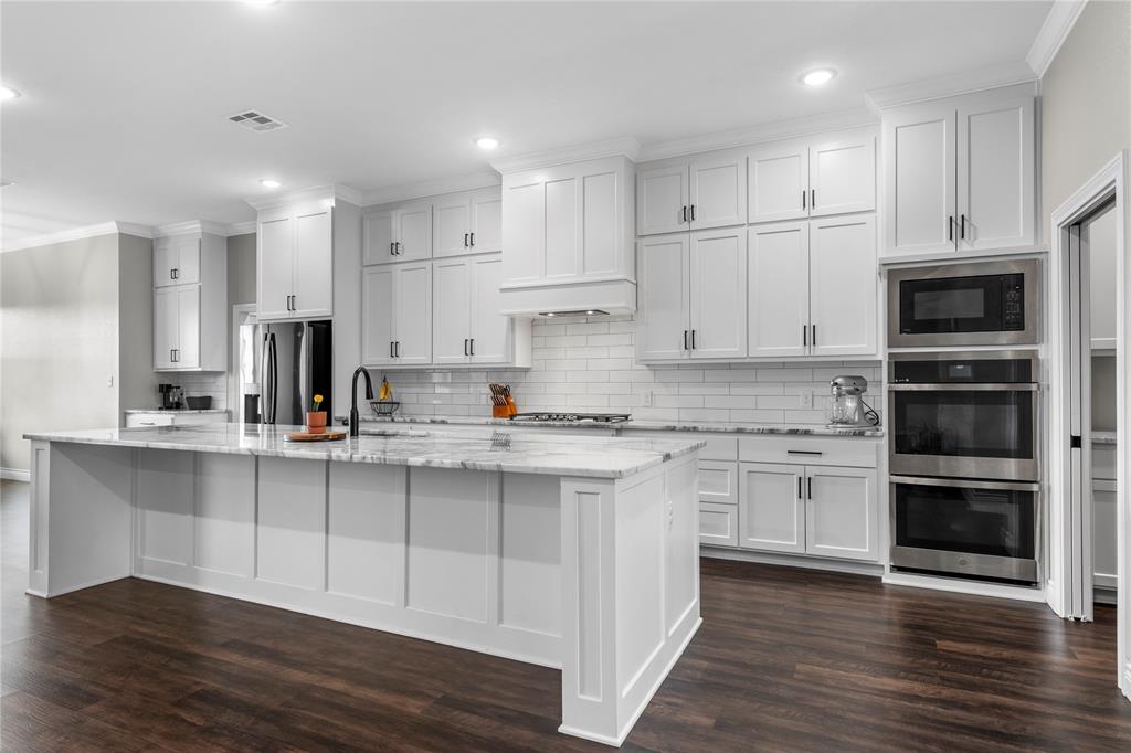 Kitchen featuring white shaker-style cabinetry, a stone-finish waterfall edge island, subway tile backsplash, stainless steel built-in double wall ovens, and wood-finish flooring