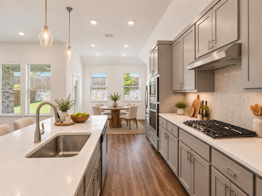 Kitchen featuring gray cabinetry, light-toned quartz countertops, a herringbone-pattern backsplash, stainless steel built-in appliances, and wood-finish flooring. Virtual photo