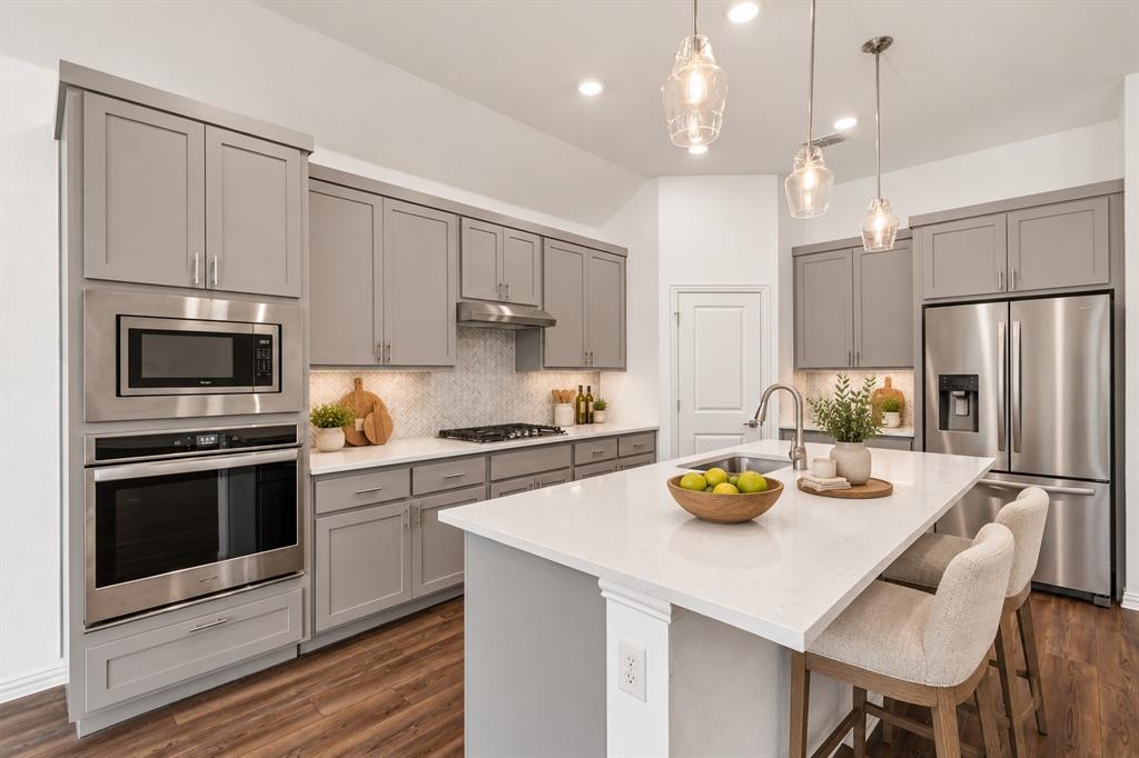 Kitchen featuring grey shaker-style cabinetry, a quartz-topped center island with a stainless steel undermount sink, herringbone-patterned tile backsplash, stainless steel built-in double oven and microwave, and wood-finish flooring. Virtual photo