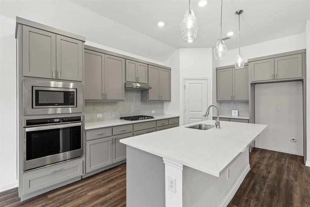 Kitchen featuring grey shaker-style cabinetry, a central island with a quartz countertop, stainless steel built-in appliances, herringbone tile backsplash, and wood-finish flooring