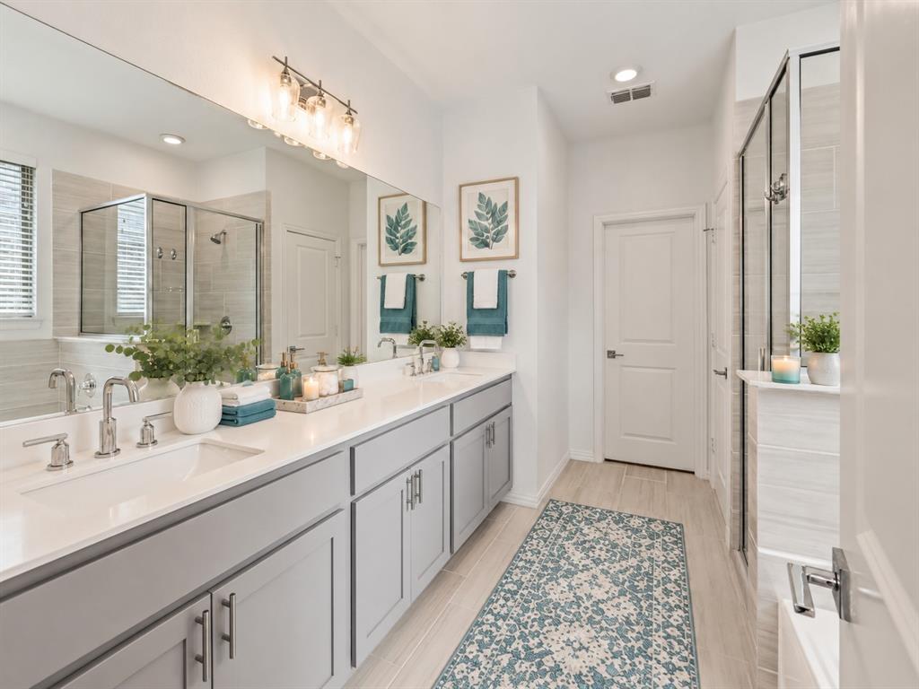 Bathroom featuring a dual-sink vanity with light gray cabinetry and white countertops, a large wall-to-wall vanity mirror, polished chrome plumbing fixtures, and a glass-enclosed shower with horizontal tile surround. Virtual photo