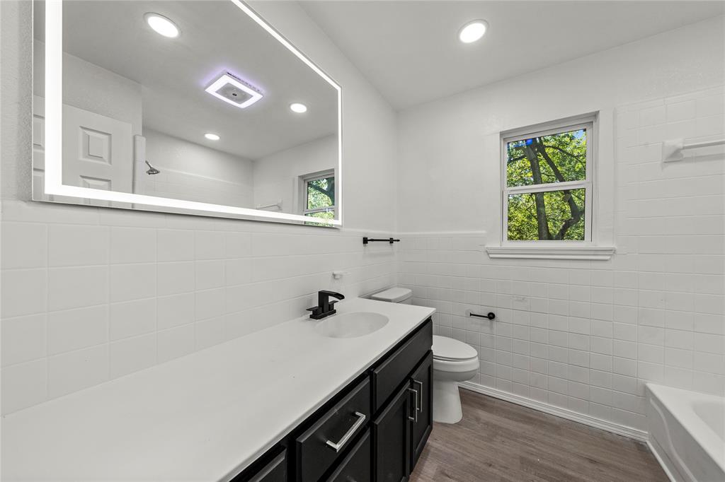 Bathroom featuring a dark wood-finish vanity with a white countertop, an integrated sink with a matte black faucet, a large rectangular LED-backlit mirror, and wood-finish flooring