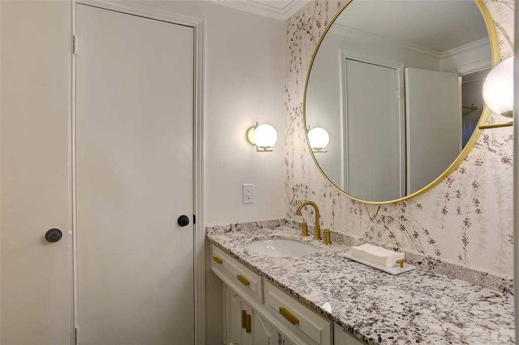 Vanity featuring an integrated sink, natural stone countertop, decorative wallpaper, circular gold-rimmed mirror, and brass-finished hardware