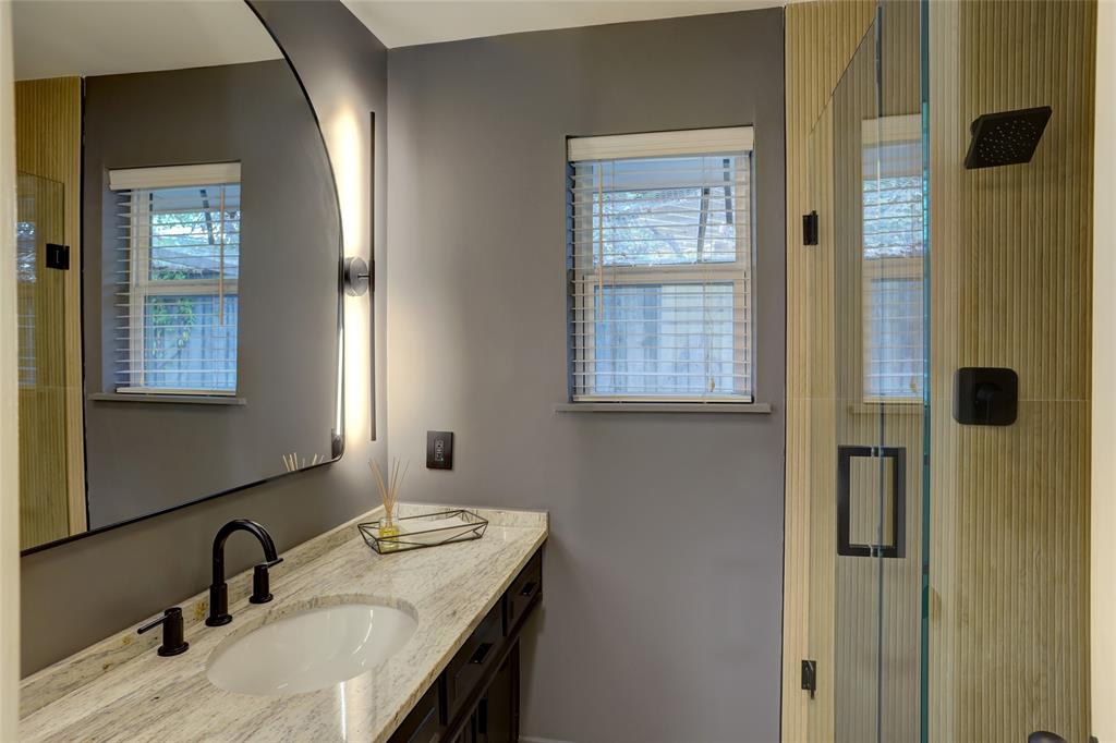 Vanity featuring a stone countertop with an integrated undermount sink, matte black widespread faucet, arched backlit mirror, and glass-enclosed shower with textured wall tile and matte black fixtures