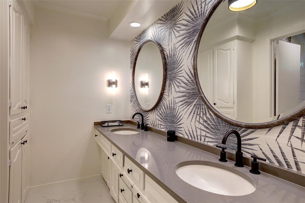 Dual-sink vanity featuring gray solid-surface countertops, white cabinetry with integrated storage, matte black widespread faucets, round mirrors, and patterned wall covering