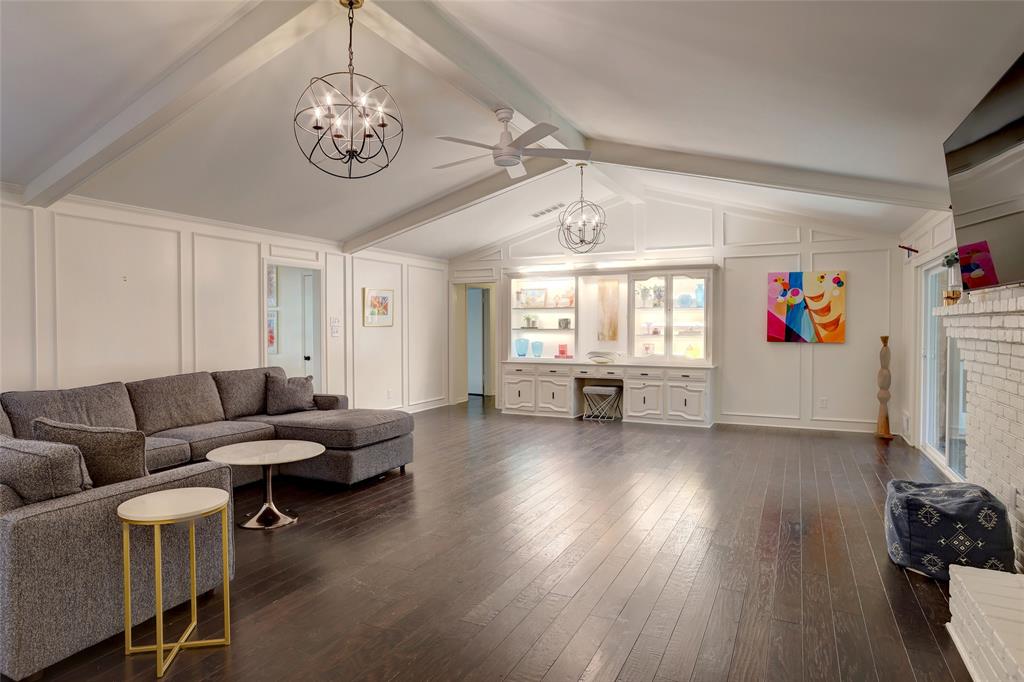 Vaulted ceiling with exposed beams, wood-finish flooring, built-in cabinetry with glass-front shelving, and a painted brick fireplace surround