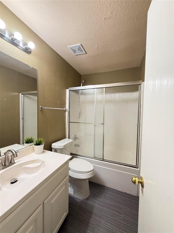 Bathroom featuring a single-sink vanity with a solid-surface countertop, full-width vanity mirror, polished chrome light fixture, and tub-shower combination with a sliding glass door and wood-finish tile flooring