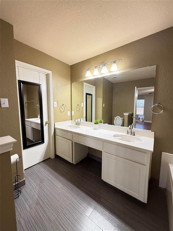 Dual-sink vanity with white cabinetry, light-toned countertop, large wall-mounted mirror, four-light vanity fixture, and wood-finish flooring