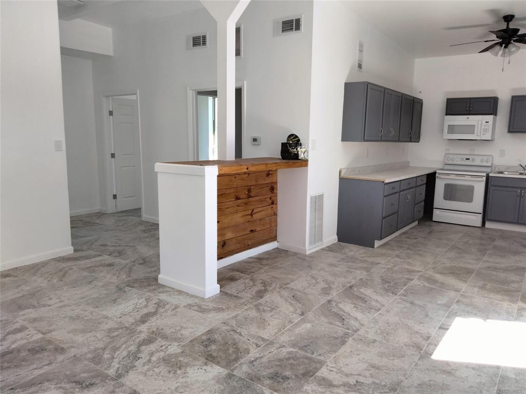 Kitchen featuring gray cabinetry, neutral solid-surface countertops, built-in white appliances, and a wood-accented breakfast bar, all complemented by large-format neutral-tone tile flooring