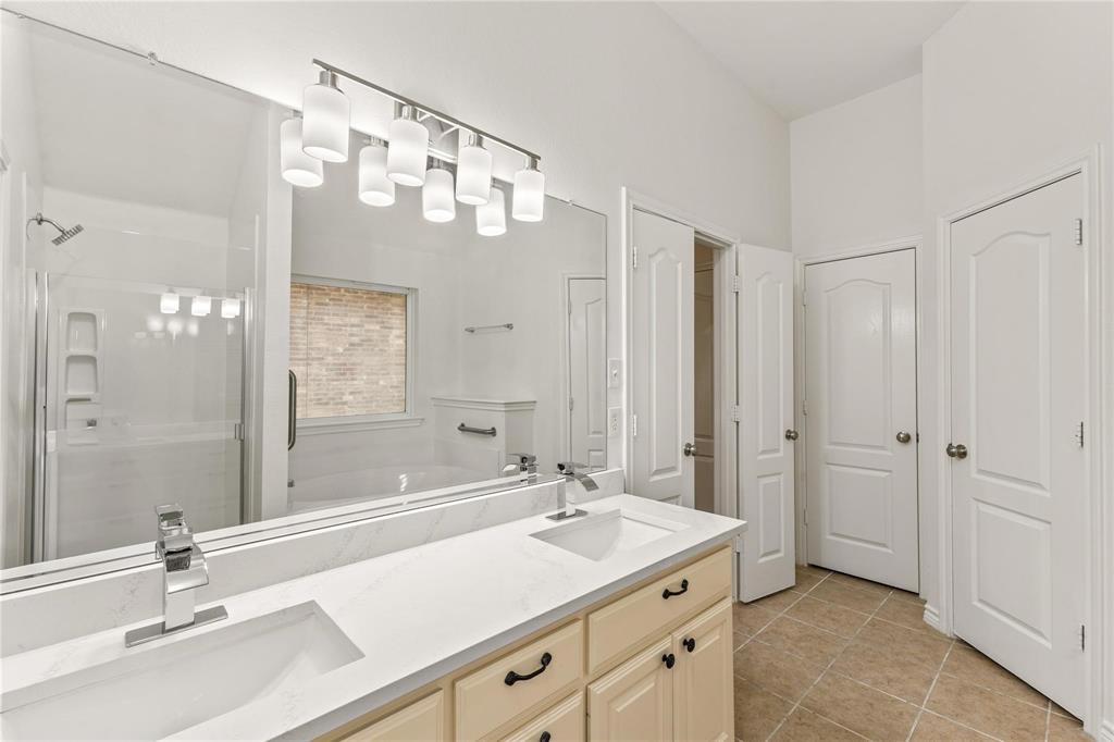 Bathroom featuring a dual-sink vanity with a solid-surface countertop, full-width wall mirror, tile flooring, and a glass-enclosed shower