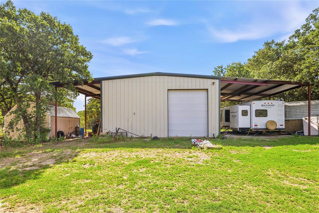 Metal-sided workshop featuring a roll-up garage door, expansive side-covered storage, and a detached secondary storage shed situated on a grassy lot with mature trees