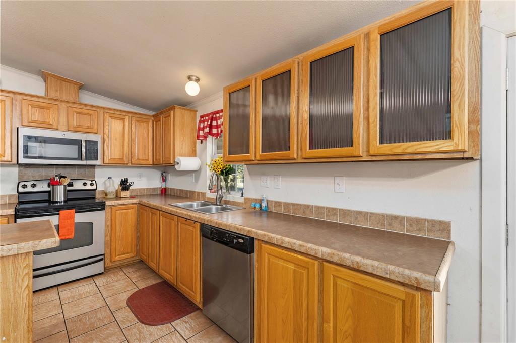 Kitchen featuring natural wood cabinetry with upper glass-panel doors, neutral stone-finish countertops, tile backsplash, and stainless steel appliances