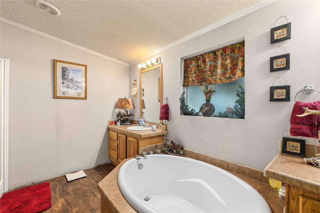 Oval soaking tub with tiled surround, wood-finish cabinetry, vanity mirror with overhead light fixture, and wood-finish flooring