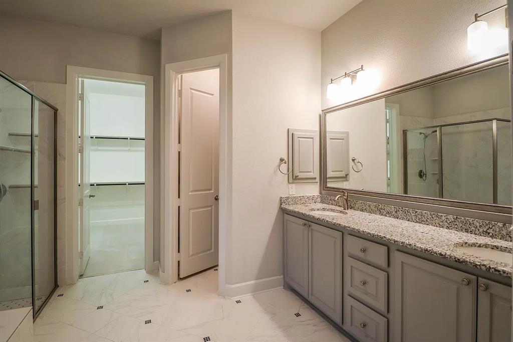 Bathroom featuring a dual-sink vanity with stone-patterned countertop, grey shaker-style cabinetry, framed vanity mirror, glass-enclosed shower, and large-format tile flooring