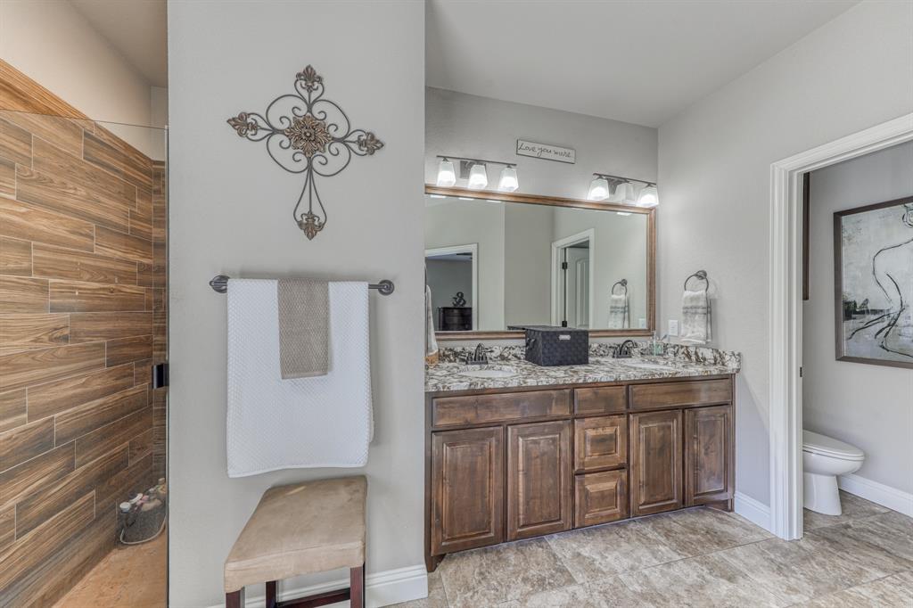 Double vanity featuring stained wood cabinetry, stone-patterned countertops, dual undermount sinks, and full-width wall mirror, complemented by a walk-in shower with wood-finish tile wall surfacing
