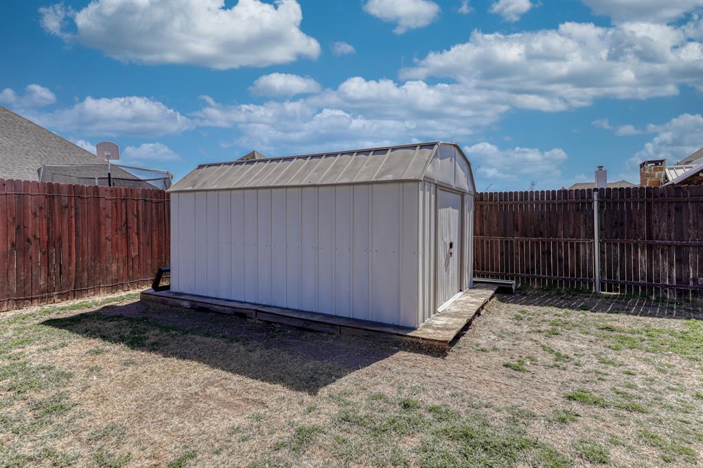 Detached metal storage shed featuring a gambrel roof and foundation-mounted platform, enclosed by a full-perimeter wood privacy fence