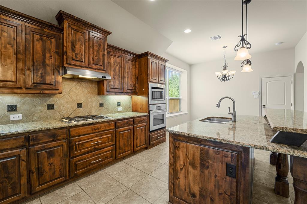 Kitchen featuring stained wood cabinetry, stone-surface countertops, tile backsplash, built-in double wall ovens, and a center island with a stainless steel sink