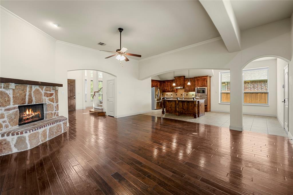 Open-concept interior featuring wood-finish flooring, a stone-faced fireplace with a timber mantel, arched architectural detailing, a central ceiling fan, and a kitchen equipped with wooden cabinetry and an island