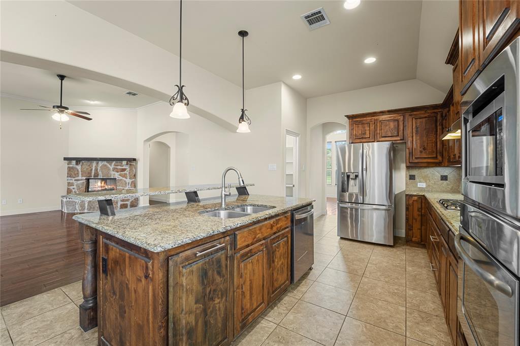 Kitchen featuring stone-finish countertops, wood cabinetry, stainless steel appliances, dual-basin sink, and tile flooring, with an additional stone-finish fireplace visible