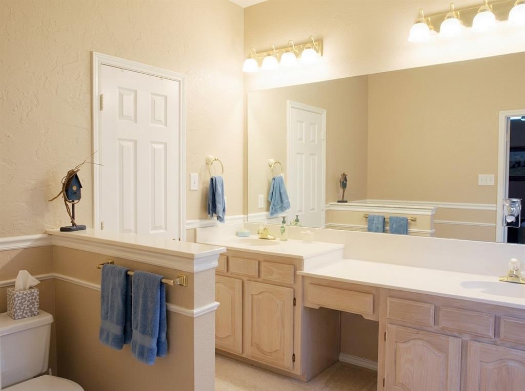 Double-sink vanity with light-tone wood cabinetry, solid-surface countertop, full-width wall mirror, and integrated lighting fixtures