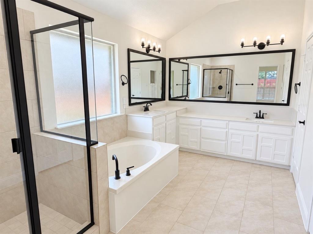 Bathroom featuring a double vanity with white cabinetry, a walk-in shower with a glass enclosure and matte black frame, a soaking tub with matte black hardware, dual framed mirrors, and tile flooring