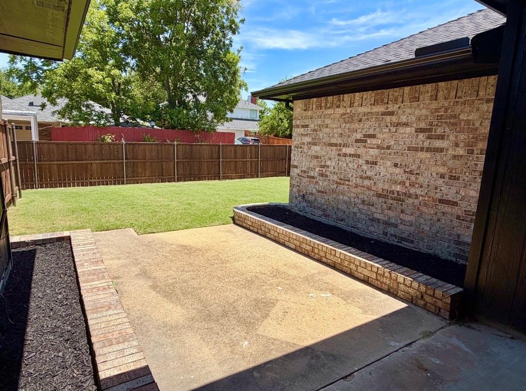 Concrete patio featuring brick-lined planting beds, brick exterior siding, and a wooden privacy fence surrounding a grass lawn
