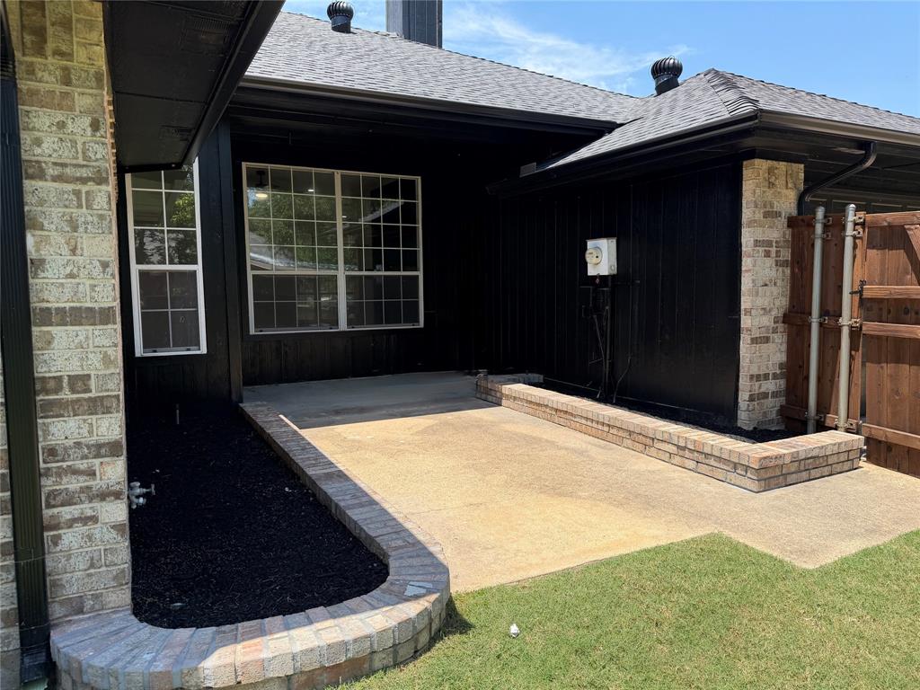 Rear exterior featuring a concrete patio, brick-trimmed landscaping beds, dark-finished vertical siding, a multi-pane grid window, and a wooden privacy fence