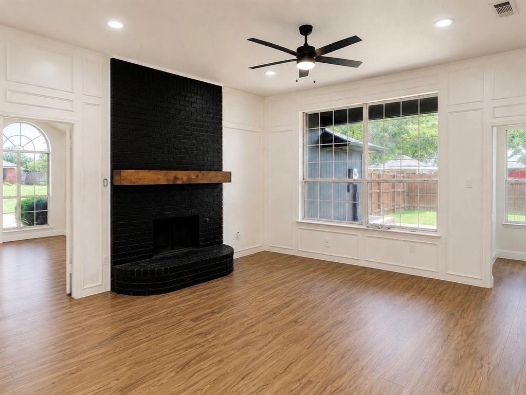 Black painted brick fireplace with a natural wood mantel, wood-finish flooring, grid-pattern windows, wainscoting wall paneling, and a ceiling-mounted light fixture with fan