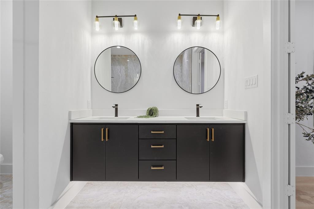 Floating dual-sink vanity with dark cabinetry, brass-tone hardware, white surface countertop, dual circular mirrors, and integrated wall-mounted light fixtures