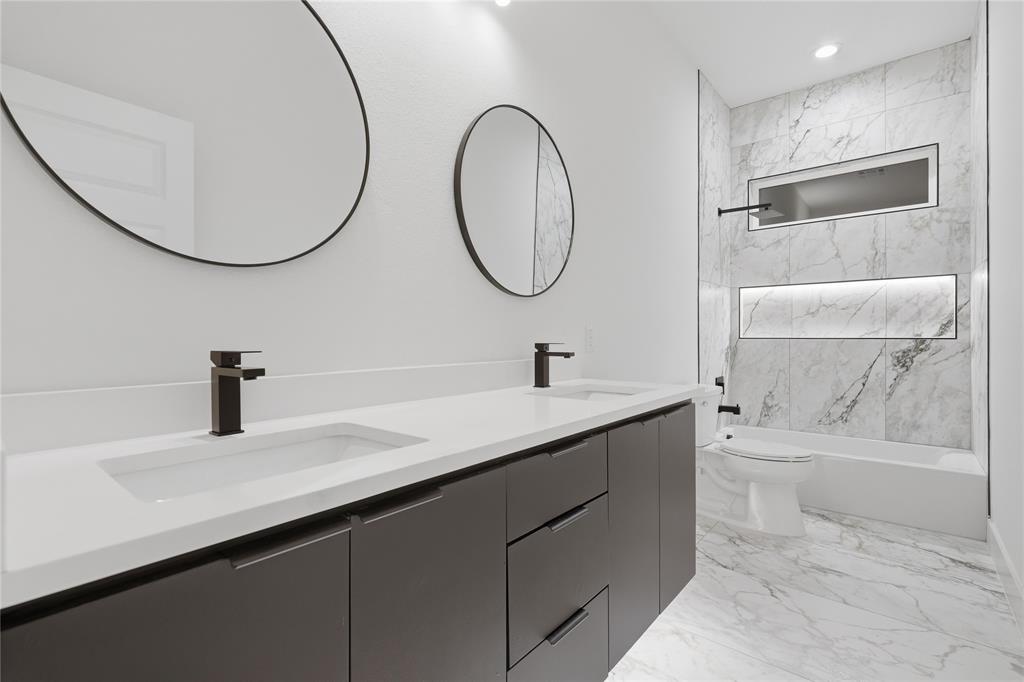 Double vanity featuring matte black faucets, circular mirrors, stone-finish tiling, and recessed wall shelving