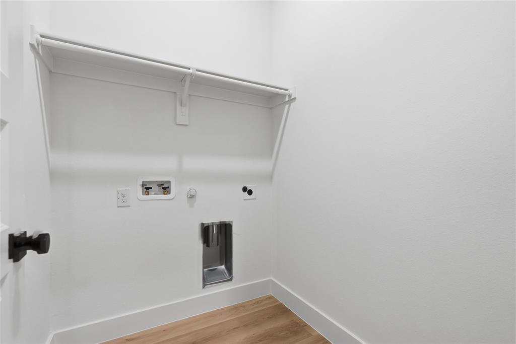 Laundry utility area featuring wood-finish flooring, a metal recessed drain box, dedicated plumbing connections, and a built-in overhead shelf with an integrated clothing rod Laundry utility area featuring wood-finish flooring, a metal recessed drain box, dedicated plumbing connections, and a built-in overhead shelf with an integrated clothing rod