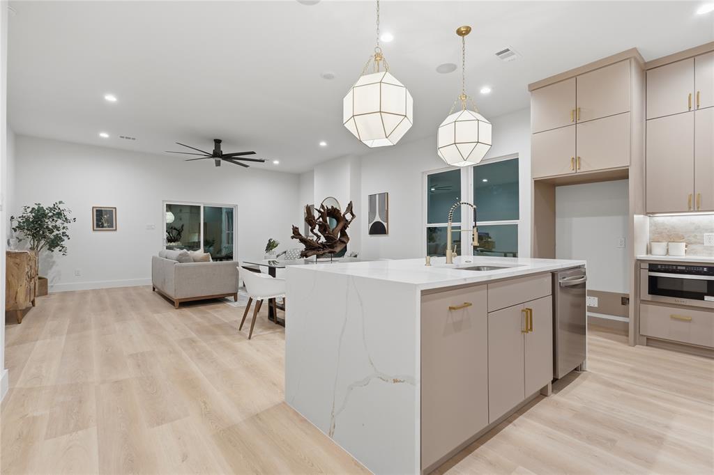 Kitchen featuring a waterfall-edge quartz island, neutral-toned cabinetry with brass hardware, stainless steel dishwasher, modern geometric pendant lighting, and wood-finish flooring Kitchen featuring a waterfall-edge quartz island, neutral-toned cabinetry with brass hardware, stainless steel dishwasher, modern geometric pendant lighting, and wood-finish flooring