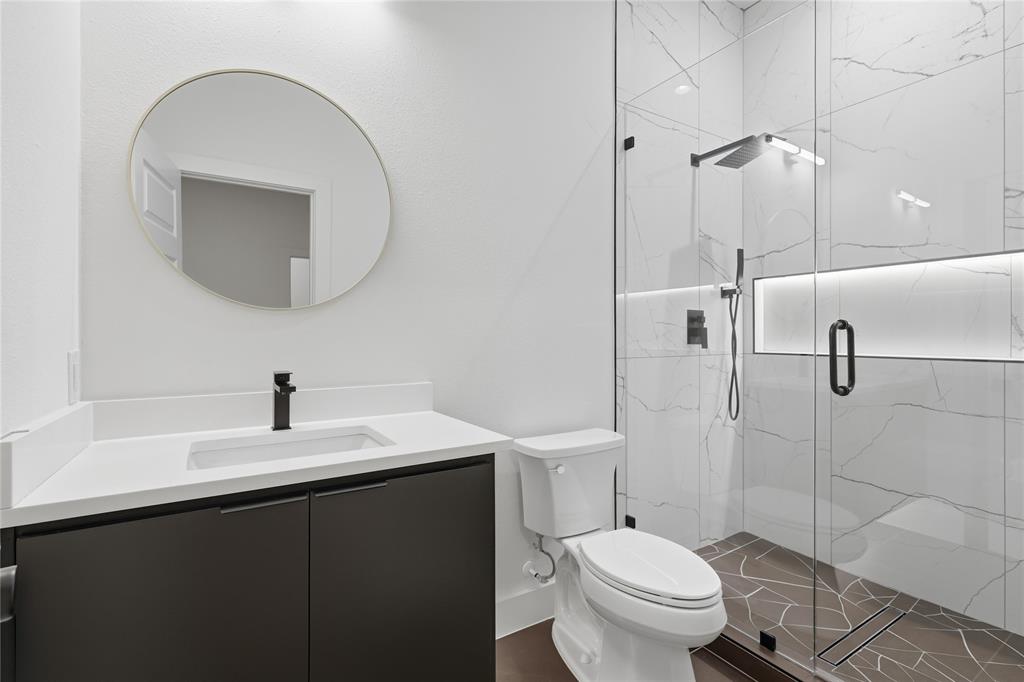 Modern bathroom featuring a dark cabinetry vanity with a white countertop, circular wall-mounted mirror, walk-in shower with large-format tile surround, integrated recessed lighting, and matte black fixtures