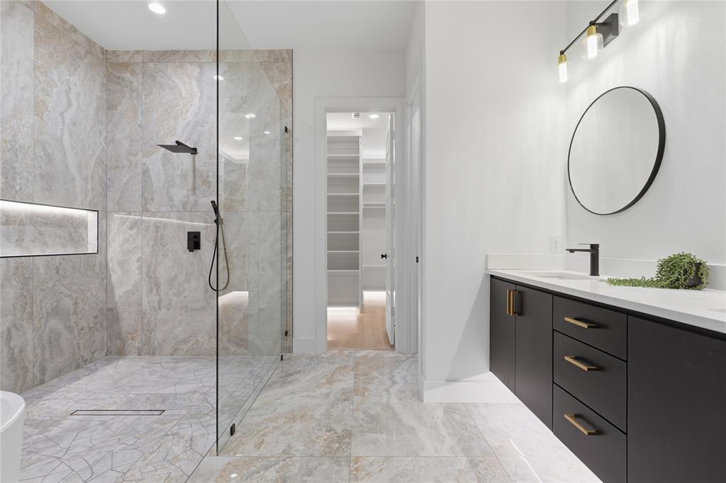 Walk-in shower with floor-to-ceiling tile, integrated recessed shelving, frameless glass enclosure, matte black rainfall shower fixture, and vanity with dark cabinetry, stone countertop, and circular mirror