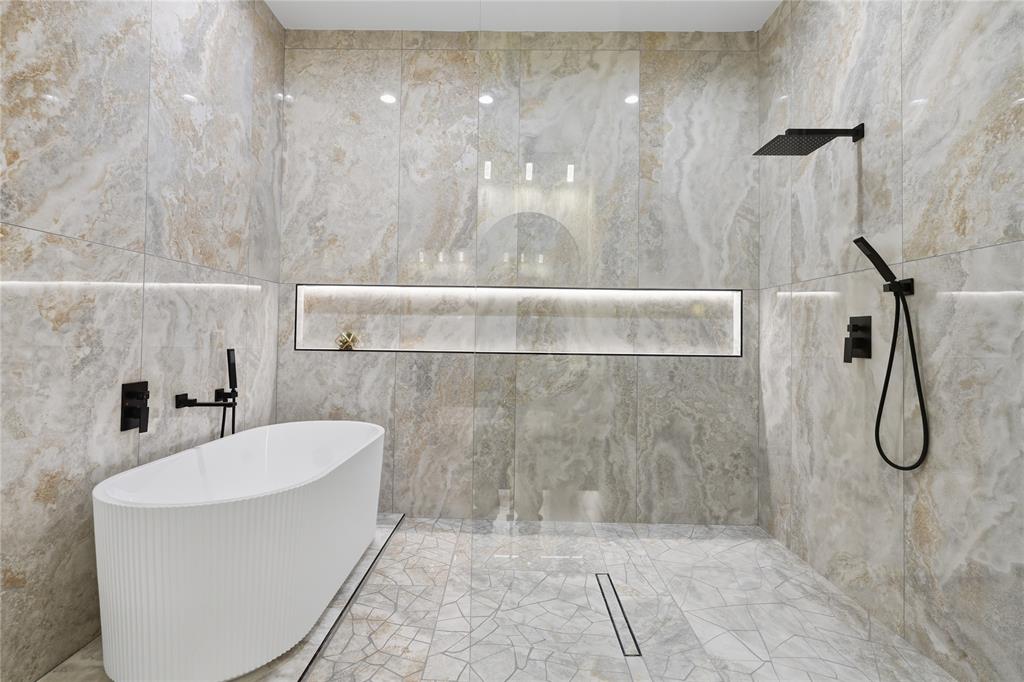 Freestanding white soaking tub, floor-to-ceiling large-format stone tile walls, matte black rainfall shower head with handheld attachment, integrated wall-recessed shelf with lighting, and linear floor drain