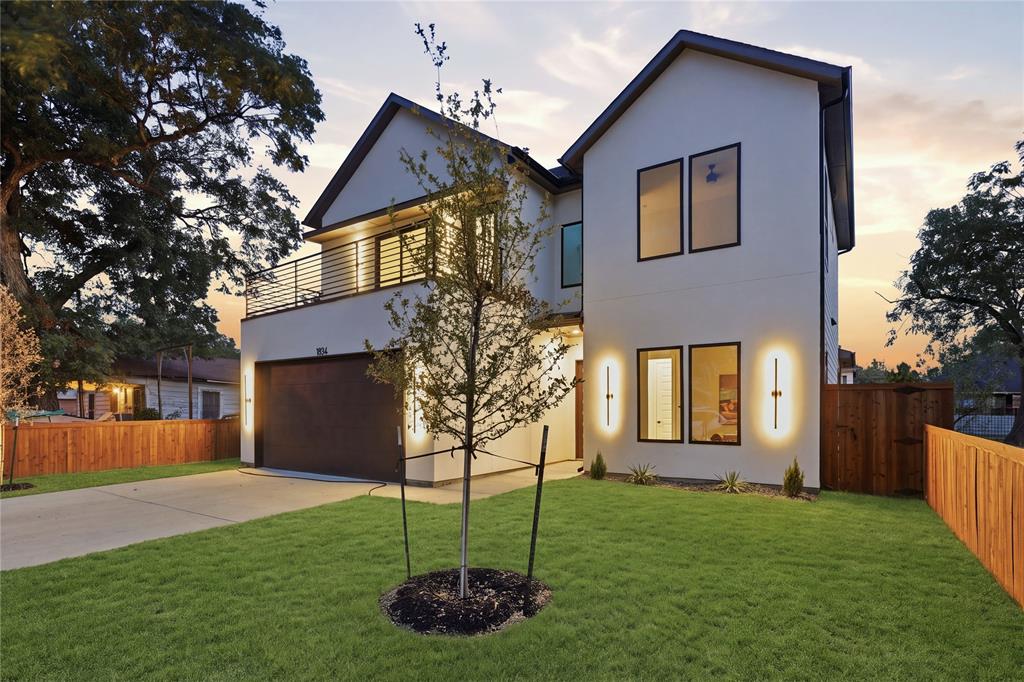 Two-story residence featuring a smooth stucco exterior, a second-level balcony with horizontal metal railing, a concrete driveway, modern wall-mounted exterior lighting, and a wood privacy fence Two-story residence featuring a smooth stucco exterior, a second-level balcony with horizontal metal railing, a concrete driveway, modern wall-mounted exterior lighting, and a wood privacy fence