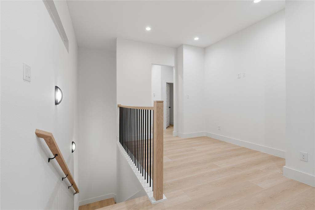Interior hallway featuring wood-finish flooring, recessed lighting, matte black wall-mounted sconces, and a staircase railing with wood handrails and black vertical balusters