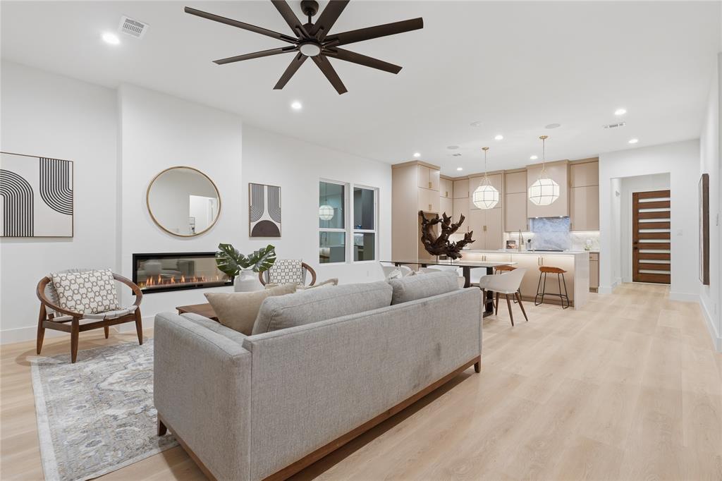 Open-concept living space featuring wood-finish flooring, a horizontal built-in fireplace, a ceiling fan with integrated lighting, recessed lighting, and kitchen cabinetry with a light stone-finish backsplash