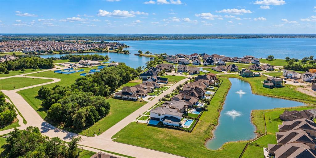 Aerial perspective of waterfront property featuring a large lake, community pond with fountains, mature trees, and paved street access