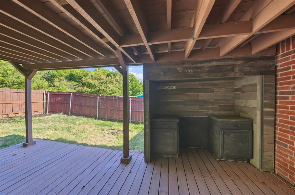 Covered wooden deck featuring an outdoor kitchen station with built-in cabinetry, wood-paneled backsplash, and brick exterior wall finish