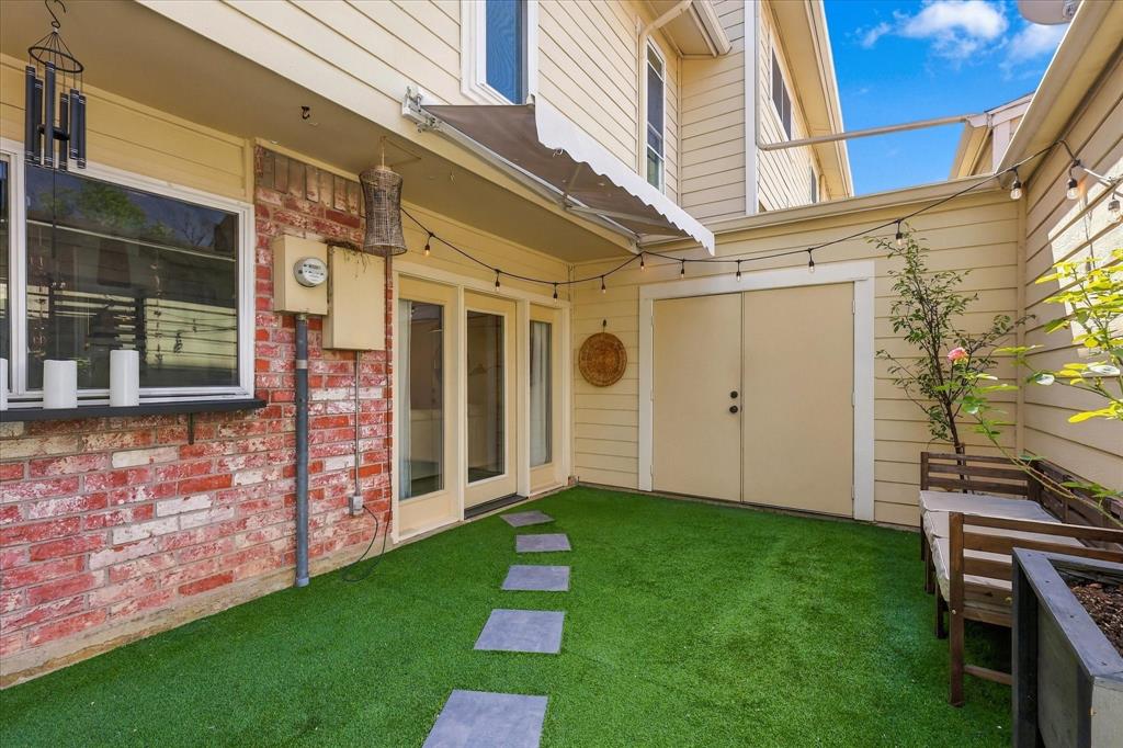 Private patio area featuring a brick facade, synthetic turf, stone paver path, retractable awning, and double-door storage