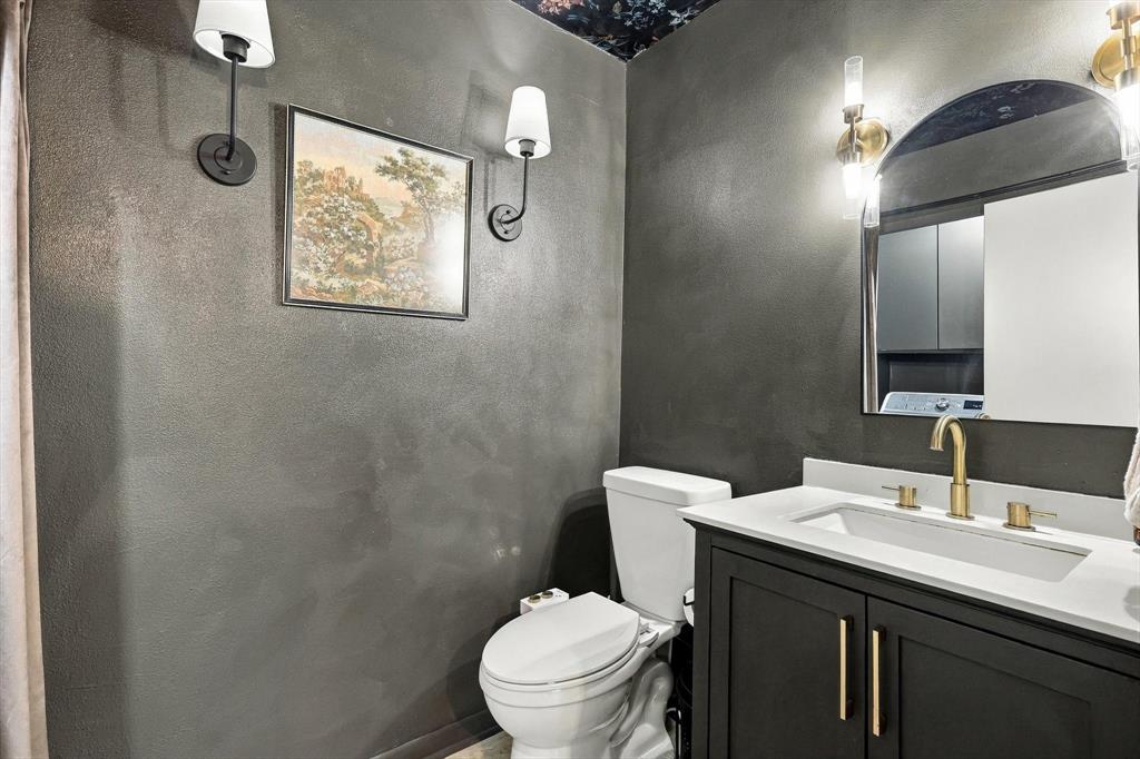 Dark vanity with a light-toned countertop, integrated basin, and brass-finish hardware, paired with dual wall-mounted light fixtures and an arched mirror