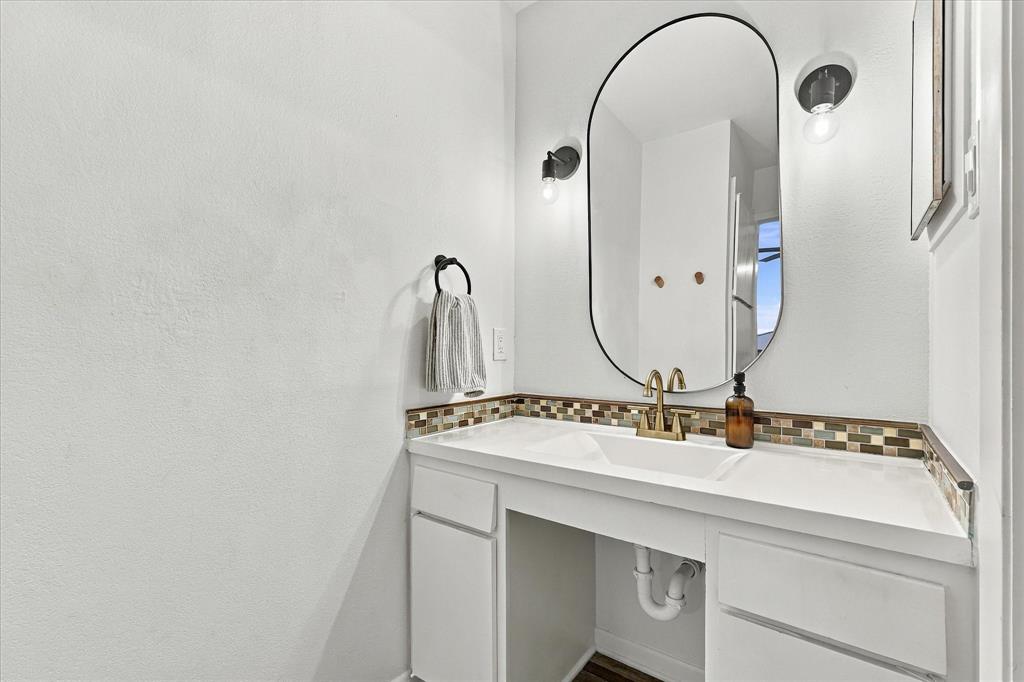 Vanity featuring an integrated sink, quartz countertop, multi-tonal mosaic tile backsplash, pill-shaped mirror, and dual wall-mounted light fixtures