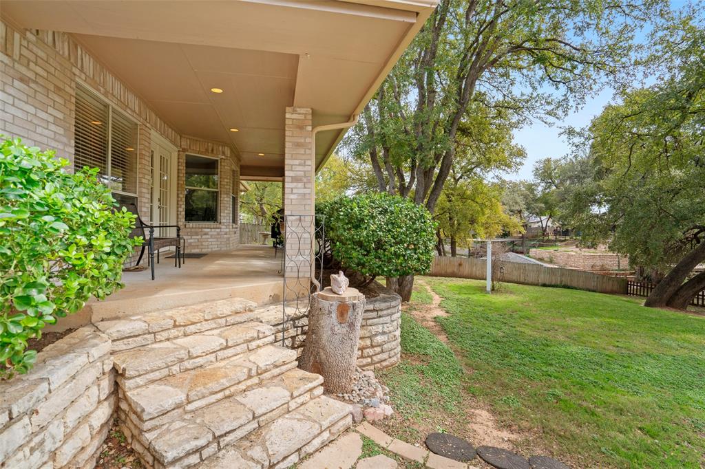 Covered porch featuring brick exterior siding, recessed lighting, and stone steps, paired with a landscaped backyard containing mature trees and stone retaining walls