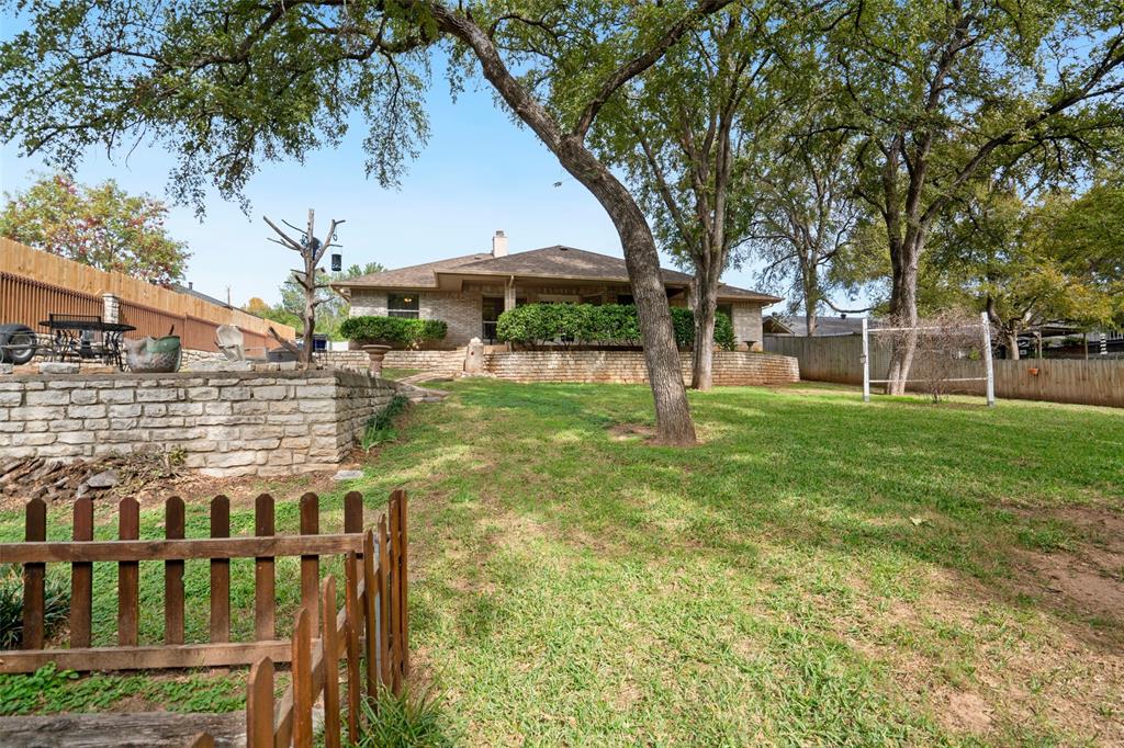 Expansive backyard featuring a tiered stone retaining wall, mature shade trees, perimeter wood fencing, and a manicured lawn