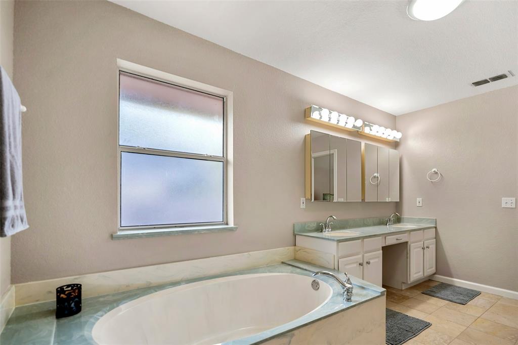 Bathroom featuring a garden tub with solid-surface surround, dual-sink vanity with built-in cabinetry, tri-fold mirrored medicine cabinet, multi-bulb vanity lighting, and ceramic tile flooring